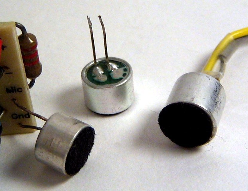 Três cápsulas de microfone de eletreto, uma delas mostrando os dois terminais soldados na traseira