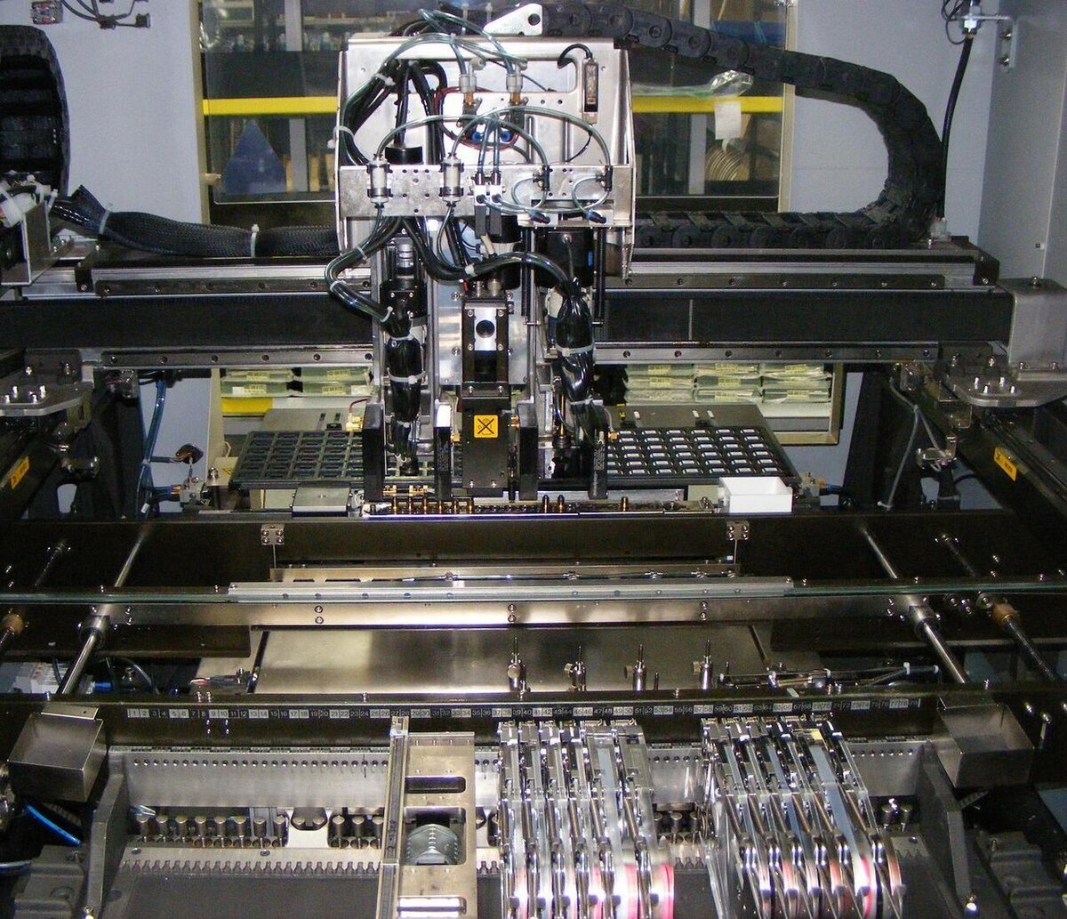 Interior de uma máquina de montagem SMD pick-and-place, com o cabeçote de bicos de vácuo, a esteira da placa e as fitas de componentes nos alimentadores