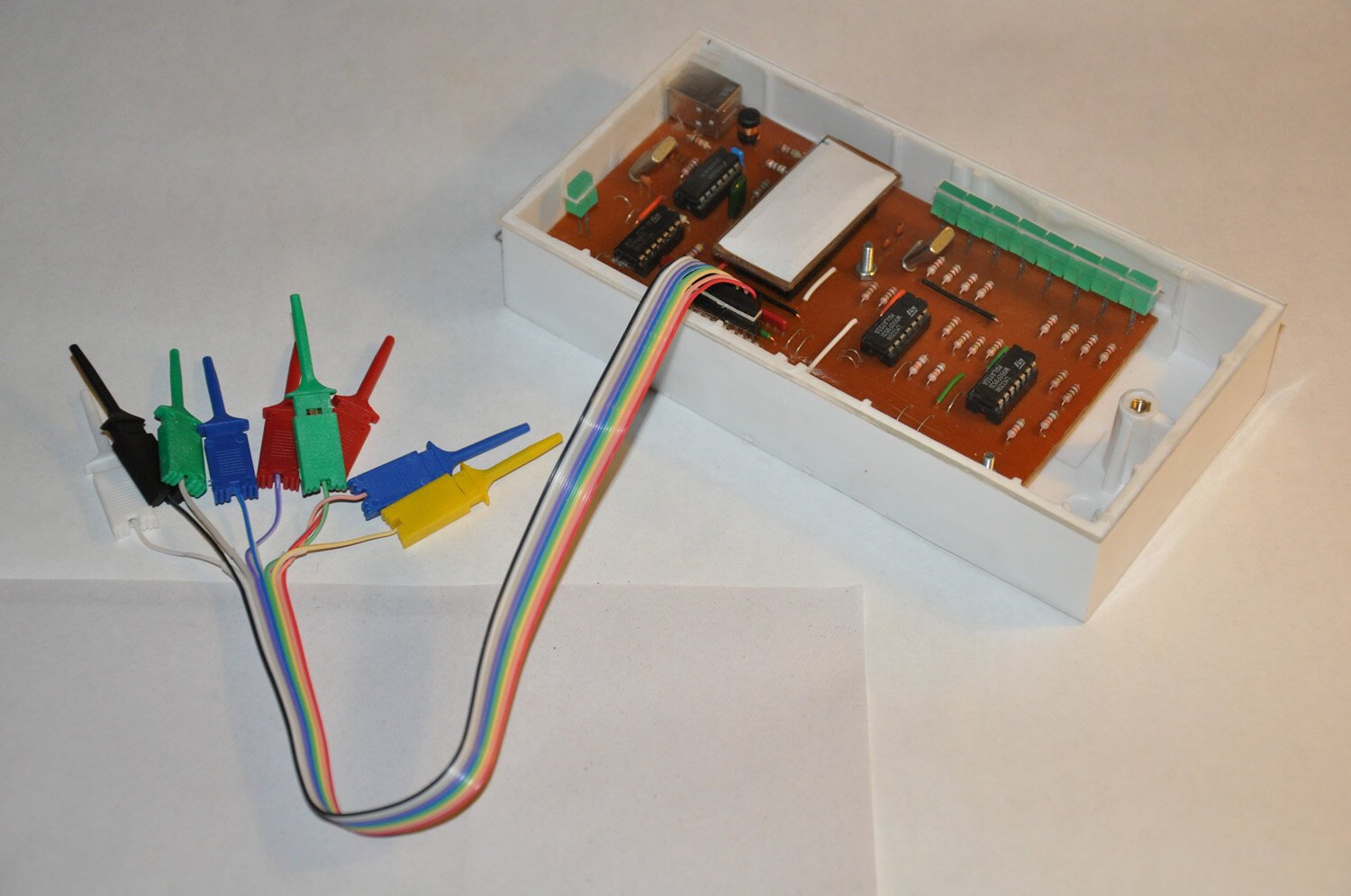 Analisador lógico USB de 8 canais com a tampa aberta mostrando a placa, ao lado do chicote de fios coloridos terminando em ganchos de teste pretos, verdes, azuis, vermelhos e amarelos