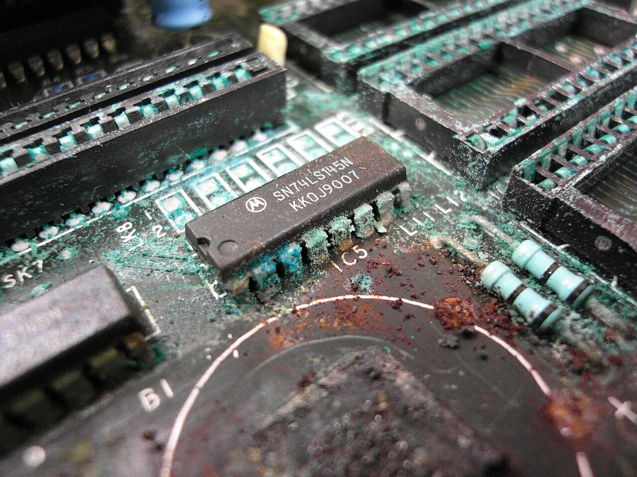 Close de placa de circuito impresso com corrosão severa: sais verdes sobre trilhas, pinos de circuitos integrados comidos e soquetes esbranquiçados